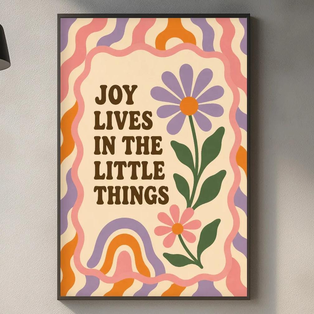 a kitchen counter with various items such as a cutting board, a soap dispenser, and a potted plant. Above the counter, there is a framed poster with the text "Joy lives in the little things" and a colorful flower design.