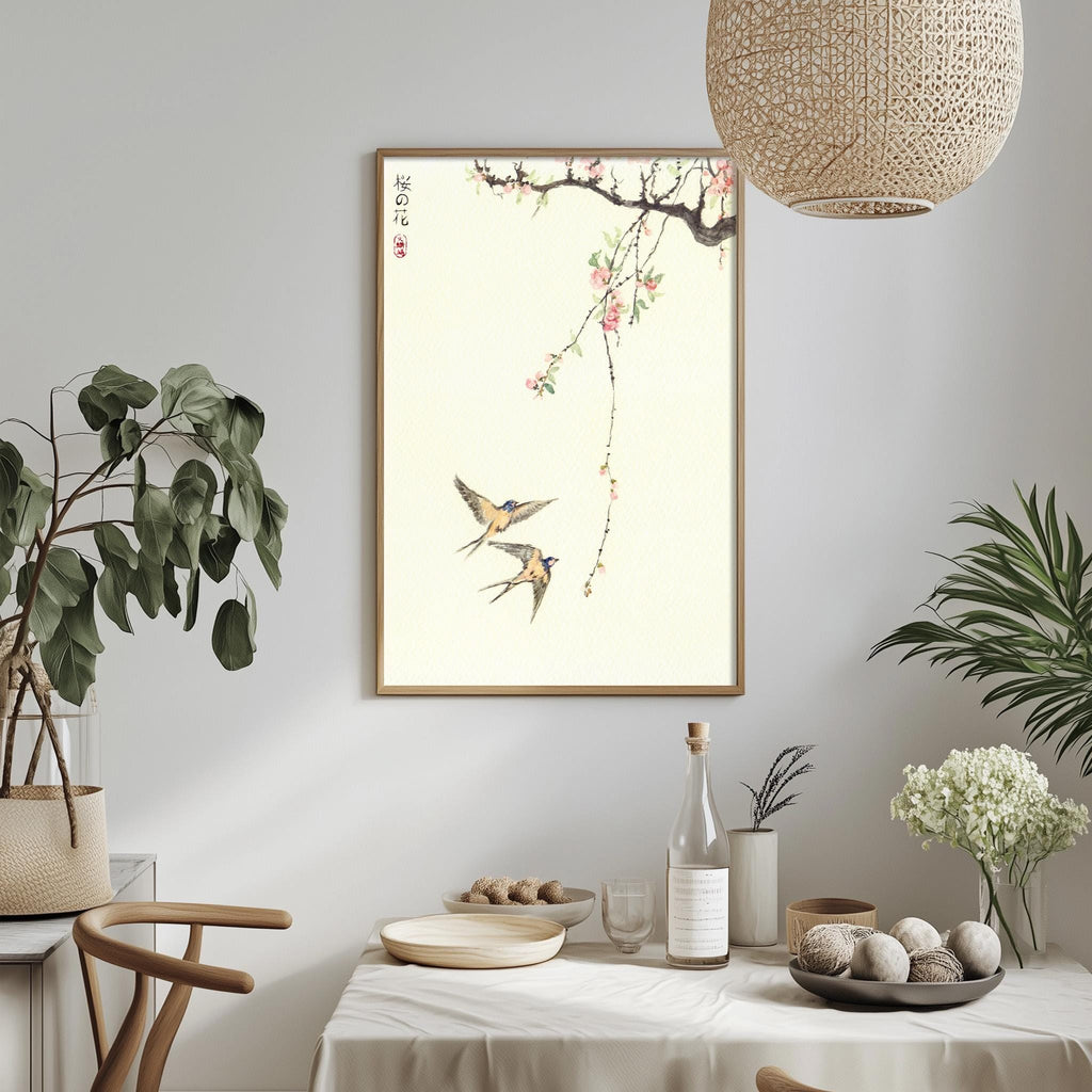 a dining table with a white tablecloth, a bowl of fruit, a bottle, and a vase with flowers. A framed picture of two birds is hanging on the wall above the table.