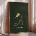 A framed poster with a green background and a yellow leaf illustration, along with the text "THRIVING" below the leaf.