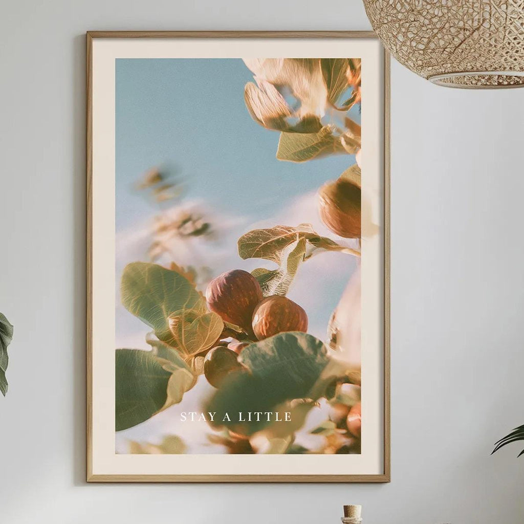 a dining table with a bowl of fruit, a bottle, and a vase of flowers, along with a framed picture of a plant hanging on the wall.