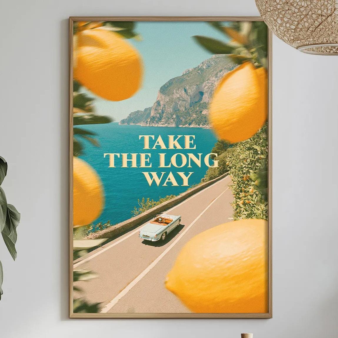 a framed poster with the text "TAKE THE LONG WAY" displayed on a wall, surrounded by a scenic view of a road with a car and mountains in the background. The poster is hung above a dining table with various items on it, including a bottle, a bowl, and a vase.