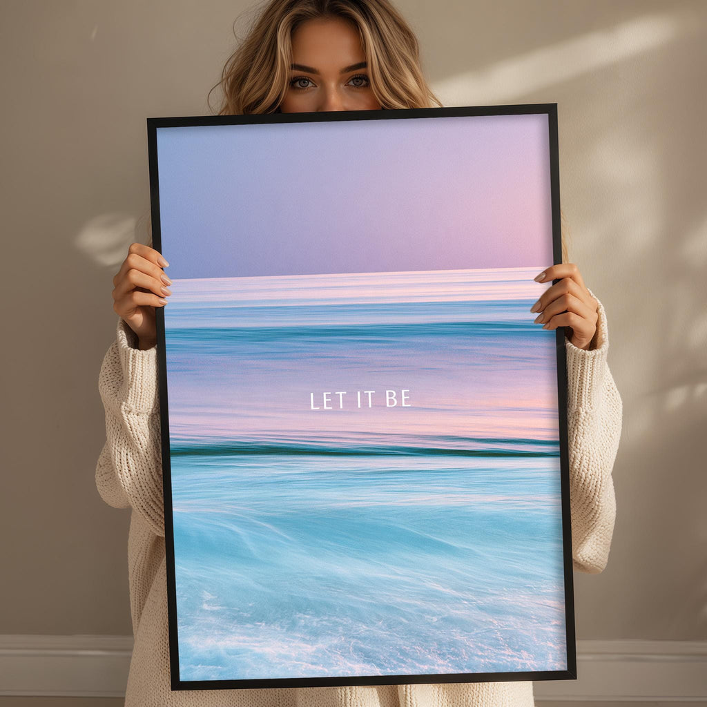 A woman holding a framed picture of a serene ocean scene with the text "LET IT BE" displayed on the picture.