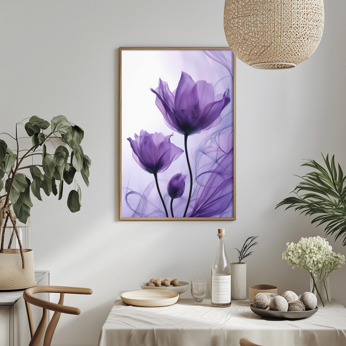 a dining table with a white tablecloth, a bowl of fruit, a bottle, and a vase with flowers. Above the table, there is a framed picture of purple flowers hanging on the wall.