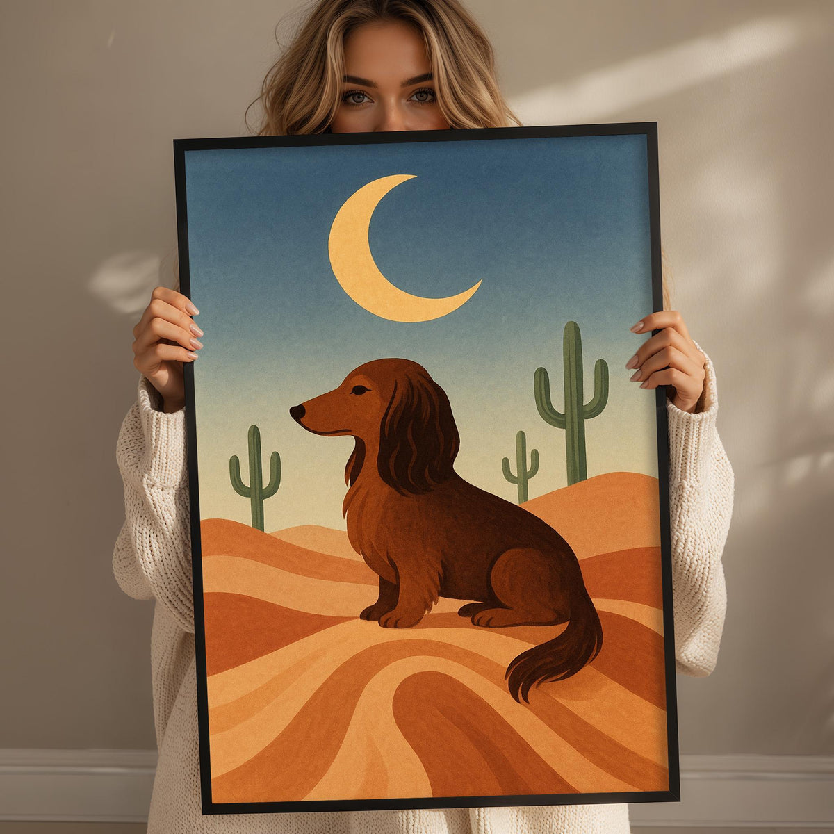 A woman holds up a framed illustration of a dachshund dog in a desert landscape, with cacti and a crescent moon in the background.