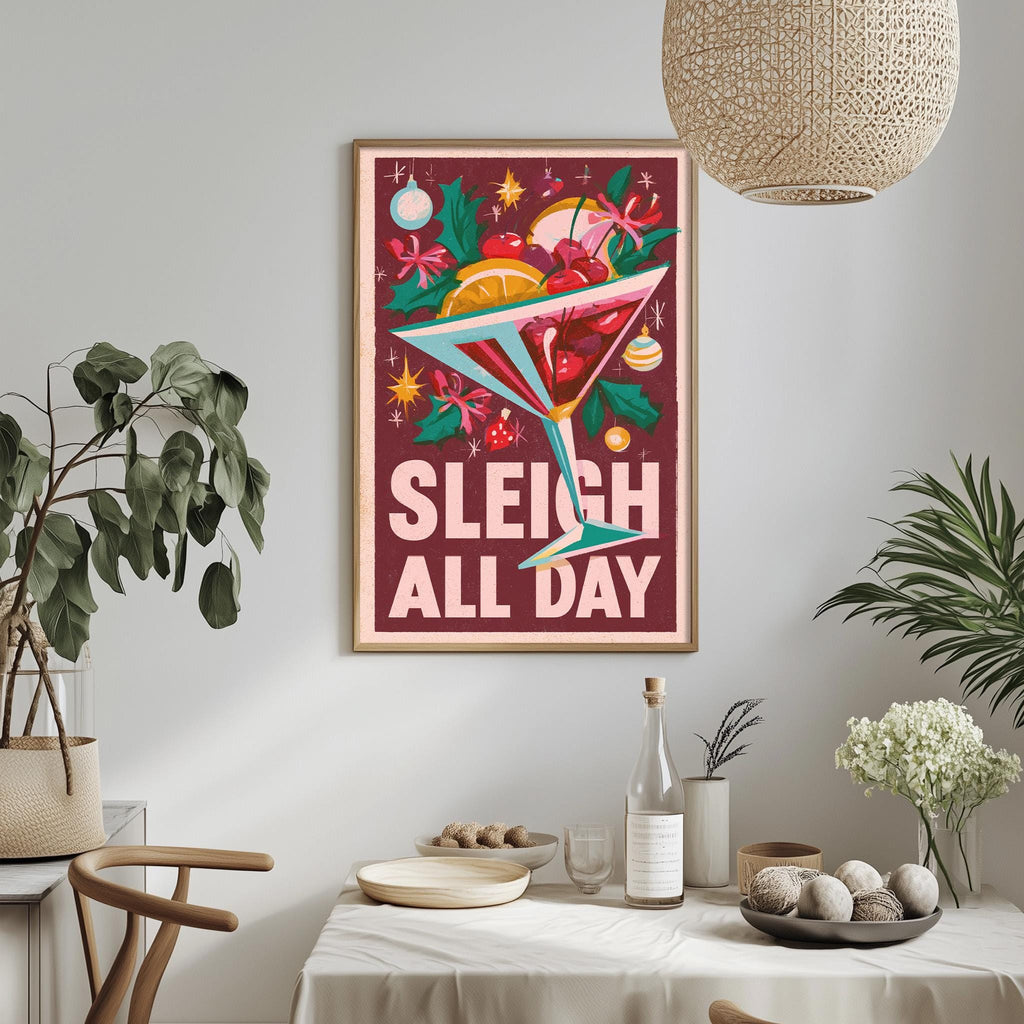 a framed poster with a colorful illustration of a cocktail glass filled with various fruits and garnishes, along with the text "Sleigh all day". The poster is hanging on a wall above a dining table, which has a vase of flowers, a bottle, and a bowl of fruit on it.