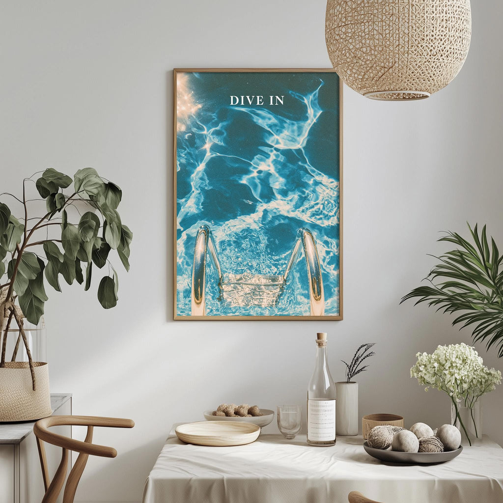 a dining table with a white tablecloth, a bottle, a bowl, and a vase with flowers. Above the table, there is a framed poster with a blue and white ocean scene, and a hanging light fixture.