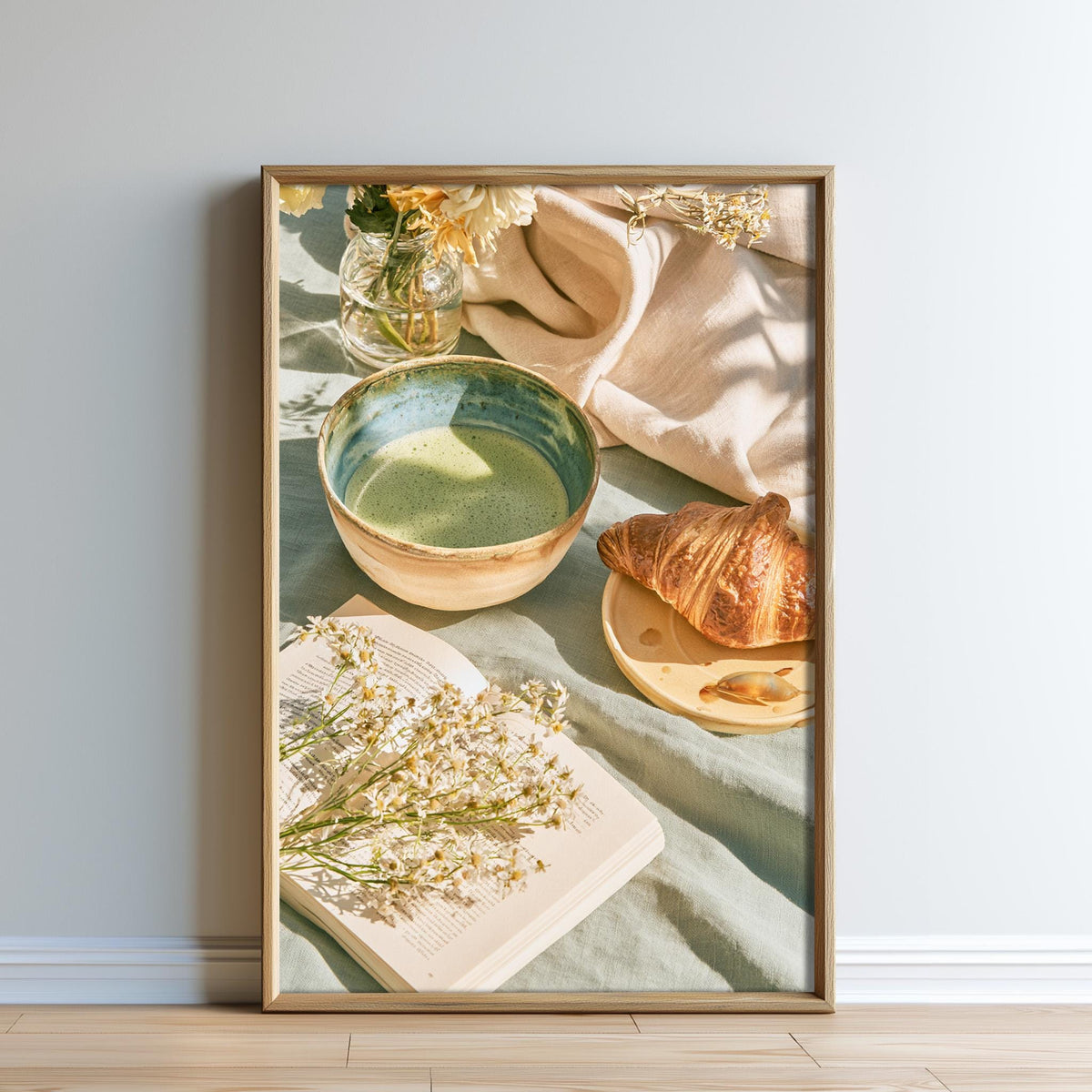 A framed image depicting a cozy breakfast scene, including a bowl of green tea, a croissant, and a bouquet of flowers.