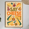 a framed poster with a vibrant design of various cheese slices and fruits, including grapes, figs, and cheese wheels, along with the text "Say Cheese" prominently displayed. The poster is hung on a wall above a dining table, which has a vase of flowers, a bottle, and a bowl of fruit on it.