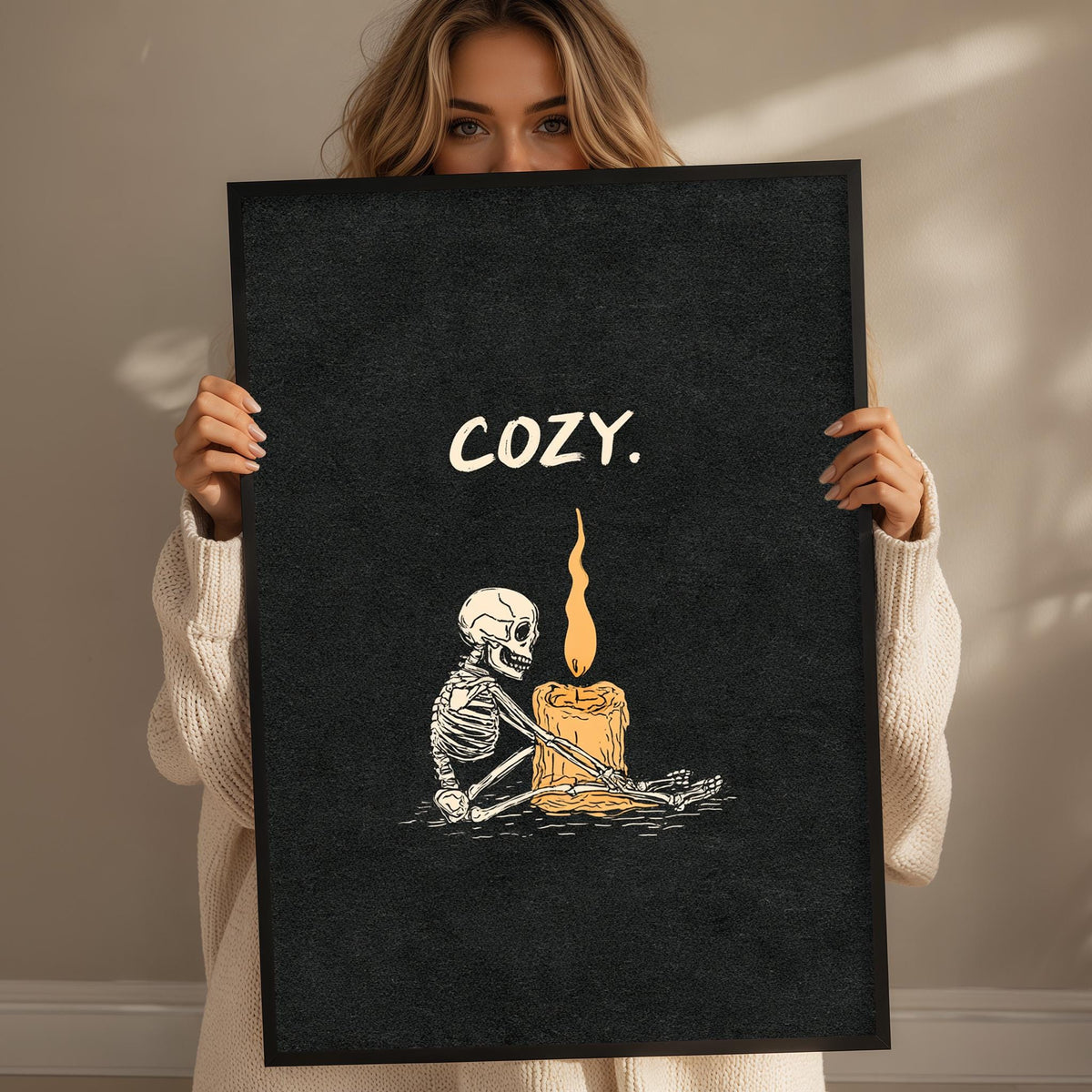 A woman holding a black poster with a skeleton and a lit candle, with the word "COZY" written on it.