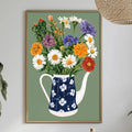 a dining table with a vase of colorful flowers, a bottle, and a bowl of fruit, all set against a white wall. Above the table, there is a framed picture of a blue and white teapot filled with flowers.