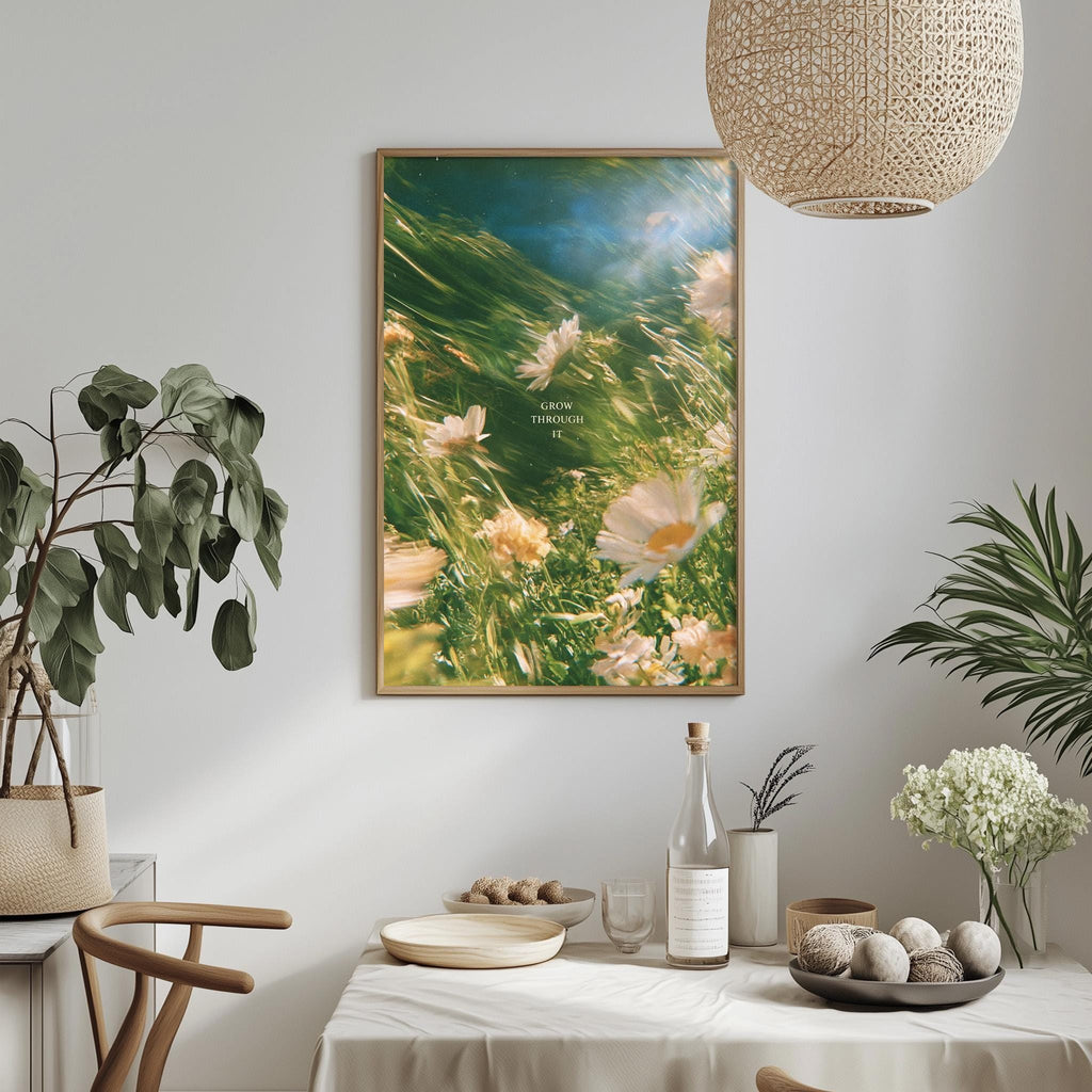 a dining table with a white tablecloth, a bowl of fruit, a bottle, and a vase with flowers. Above the table, there is a framed picture of flowers hanging on the wall.