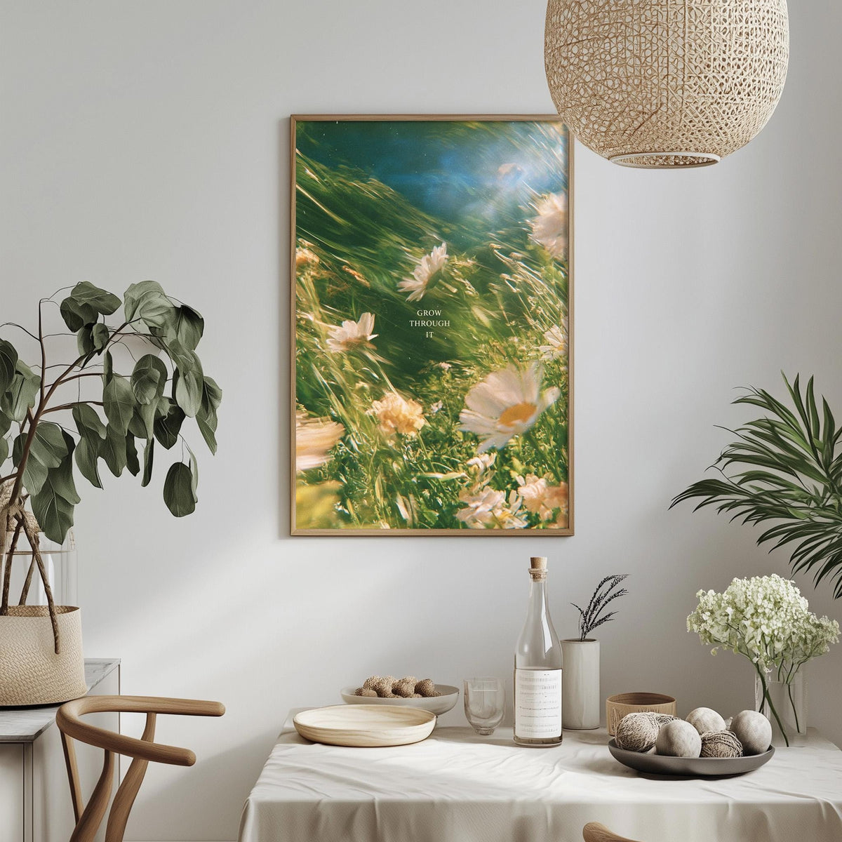 a dining table with a white tablecloth, a bowl of fruit, a bottle, and a vase with flowers. Above the table, there is a framed picture of flowers hanging on the wall.