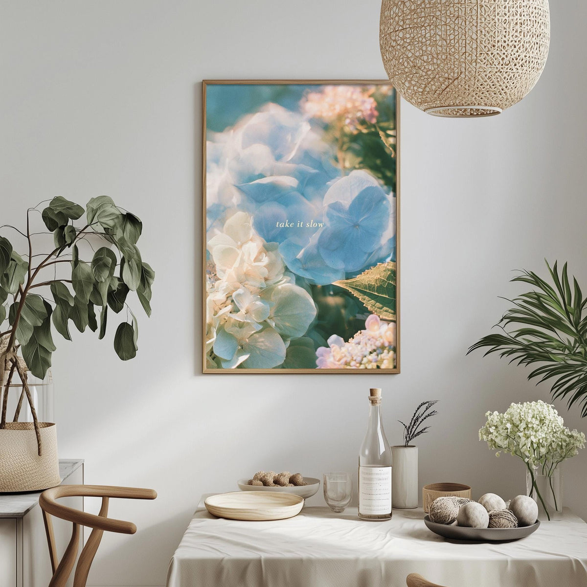 a dining table with a white tablecloth, a bowl of fruit, a bottle, and a vase with flowers. Above the table, there is a framed picture of flowers hanging on the wall.