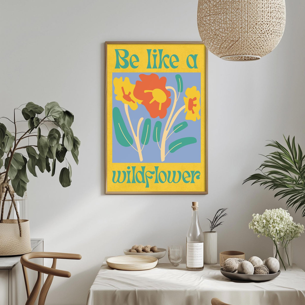 a dining table with a white tablecloth, a bowl of fruit, a bottle, and a vase with flowers. Above the table, there is a framed poster with a yellow background and a blue border, featuring a flower design and the text "Be like a wildflower".