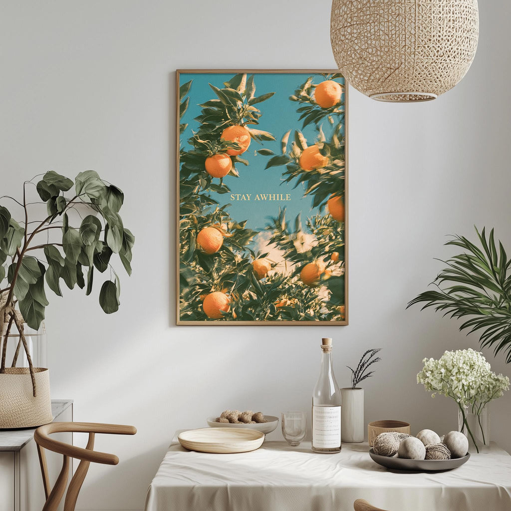 a dining table with a bowl of fruit, a bottle, and a vase of flowers, along with a potted plant and a hanging light fixture. Above the table, there is a framed poster with an orange tree and the text "STAY AWARE".