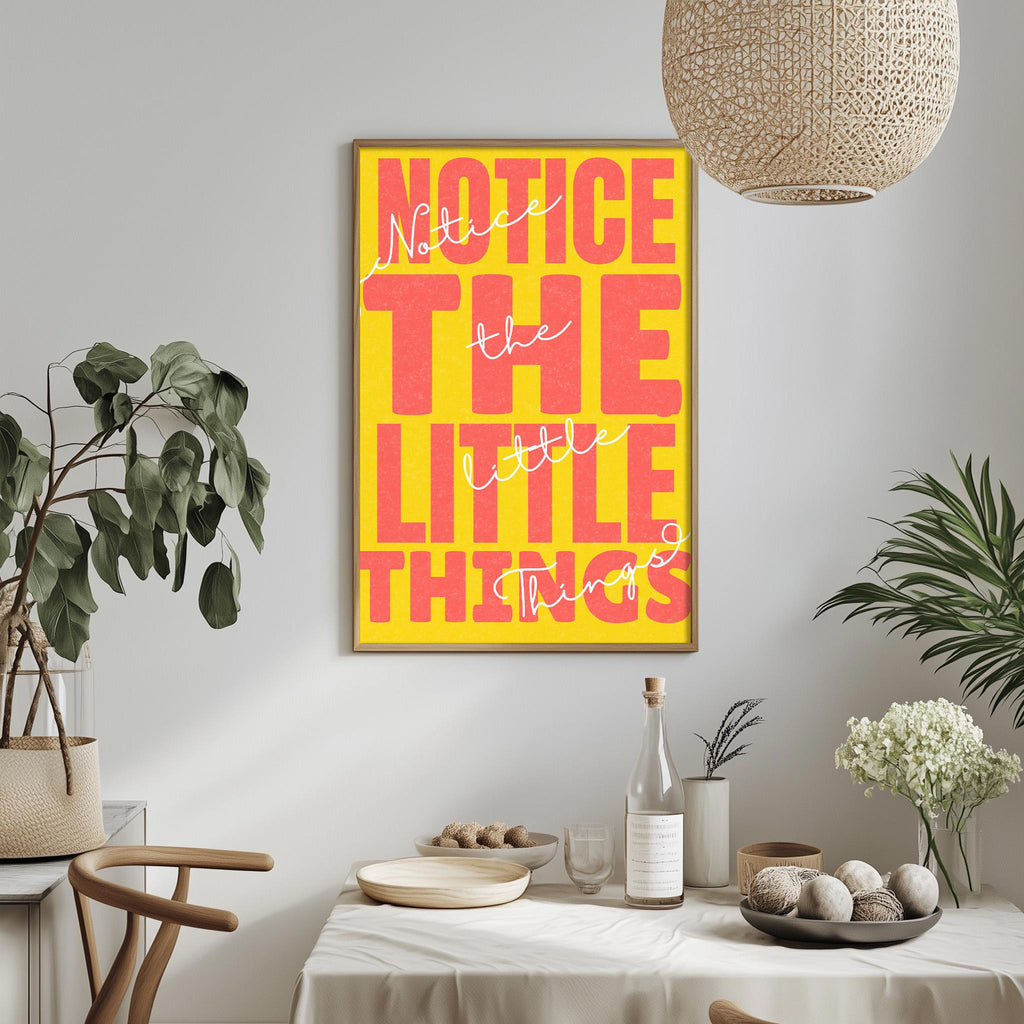 a framed poster with the text "NOTICE THE LITTLE THINGS" in a vibrant yellow and pink color scheme, hanging on a wall above a dining table with various decorative items and plants.