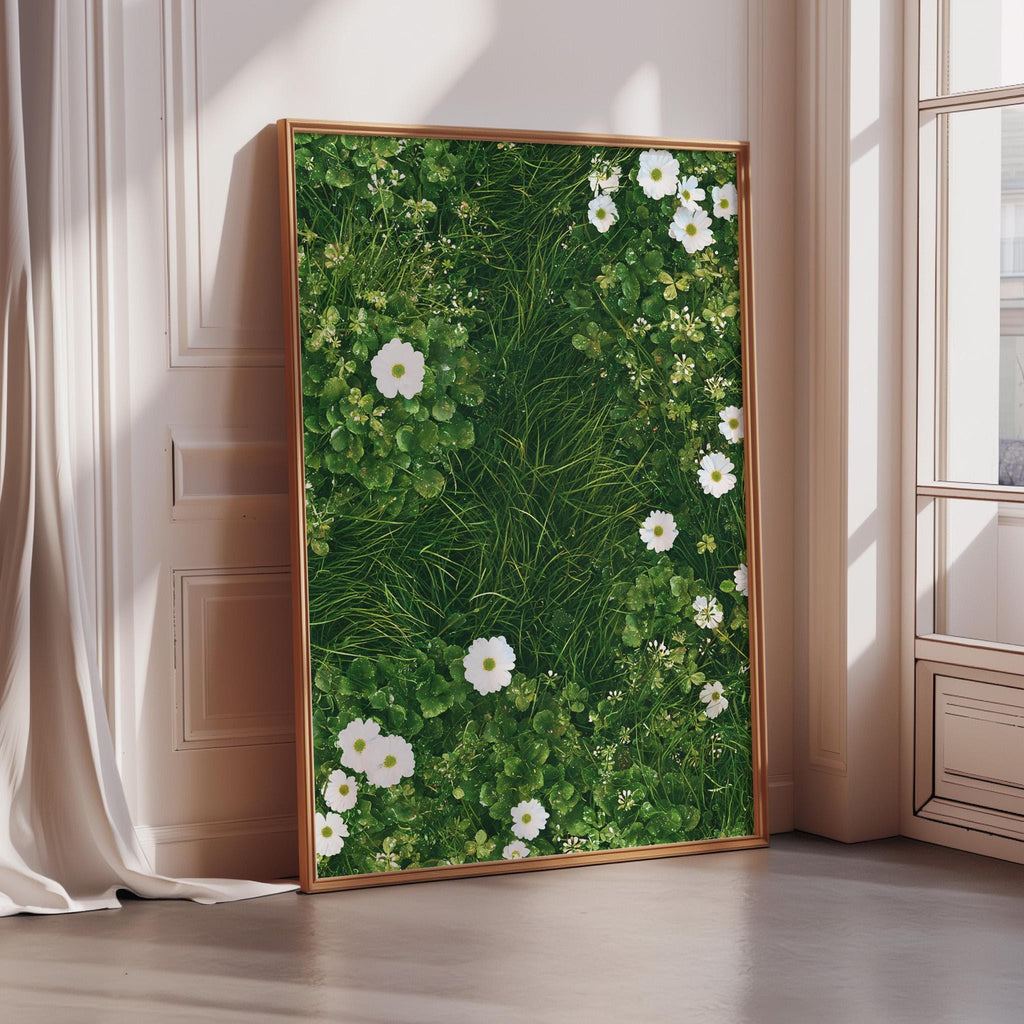 A large framed picture with a lush green grassy background and white flowers is displayed on a wall in a room with a window.