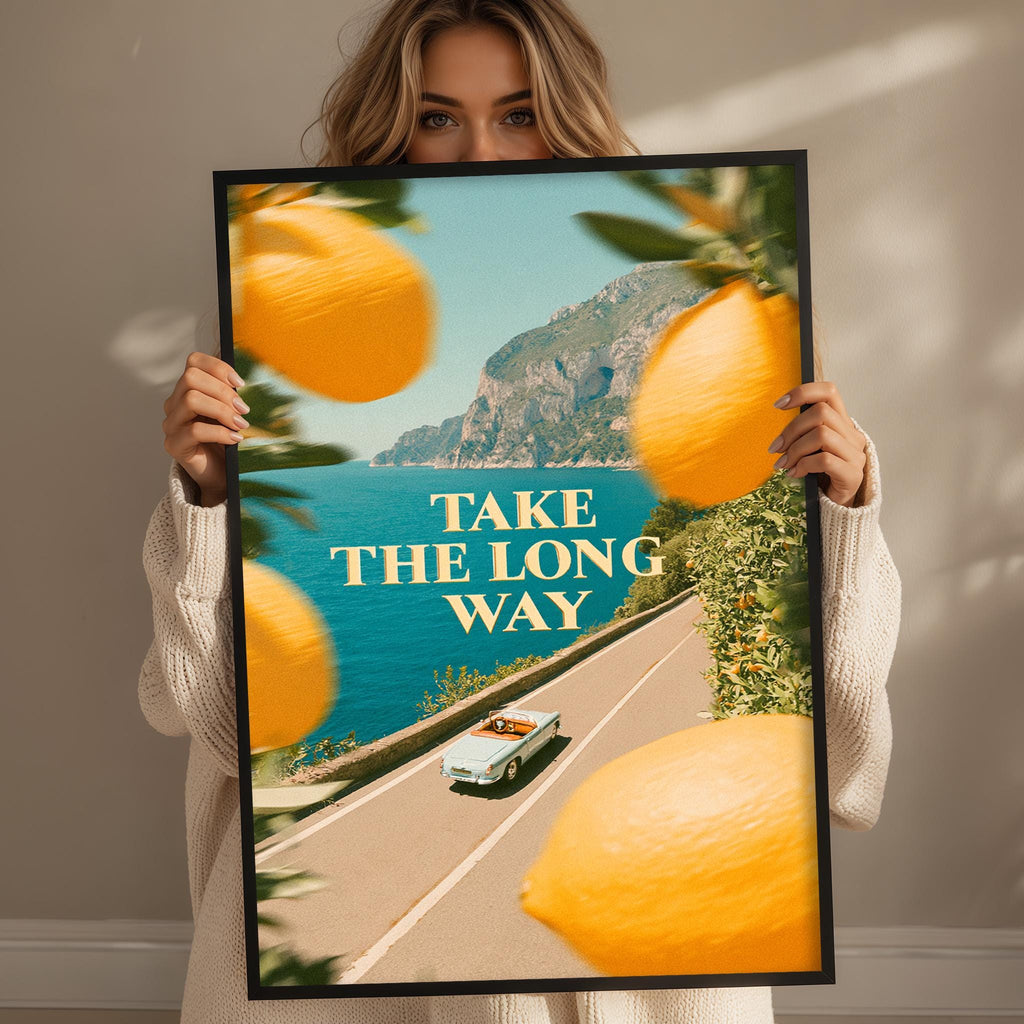 A woman holds up a framed poster with a scenic road and a car, along with the text "TAKE THE LONG WAY" displayed prominently.