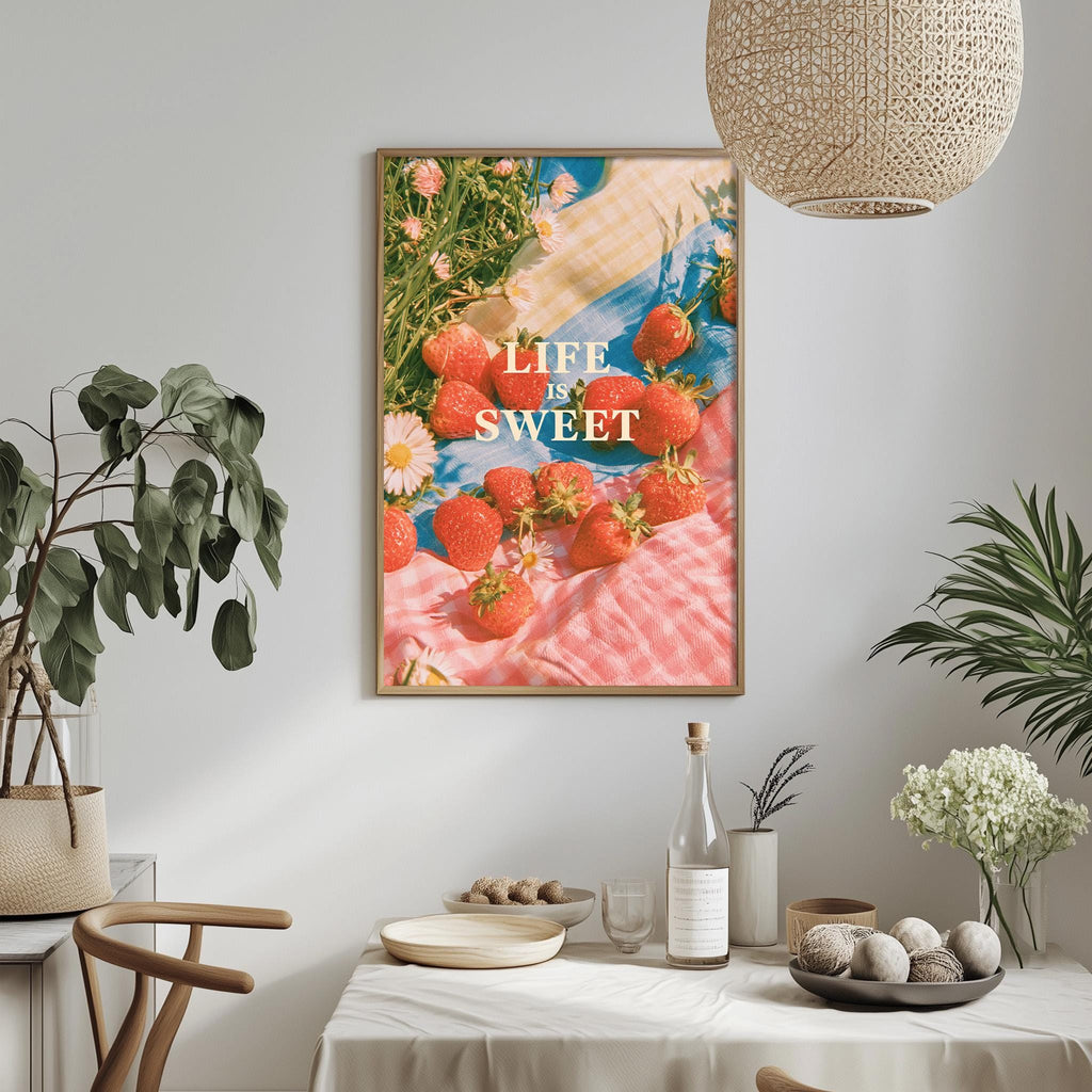 a framed poster with a vibrant image of strawberries on a pink and white checkered cloth, hanging on a wall above a dining table with various items on it.