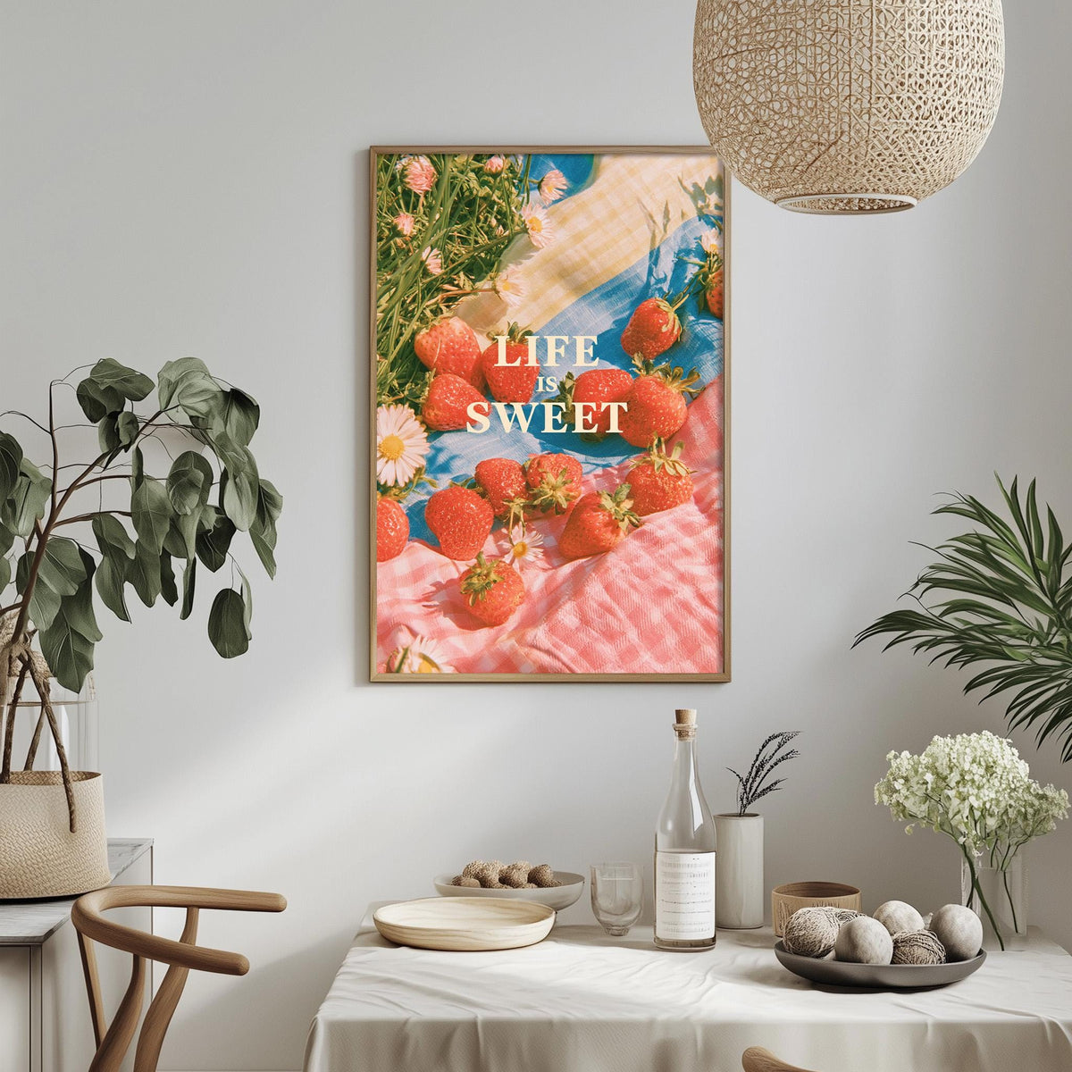 a framed poster with a vibrant image of strawberries on a pink and white checkered cloth, hanging on a wall above a dining table with various items on it.