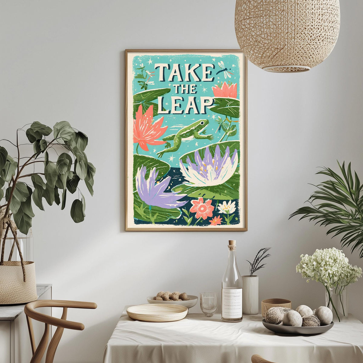 a framed poster with a vibrant illustration of a pond with colorful flowers and plants, along with the text "TAKE THE LEAP" displayed prominently. The poster is hanging on a wall above a dining table, which has various items on it, such as a bottle, a bowl, and a vase. The room also has potted plants and a hanging light fixture, creating a cozy and inviting atmosphere.