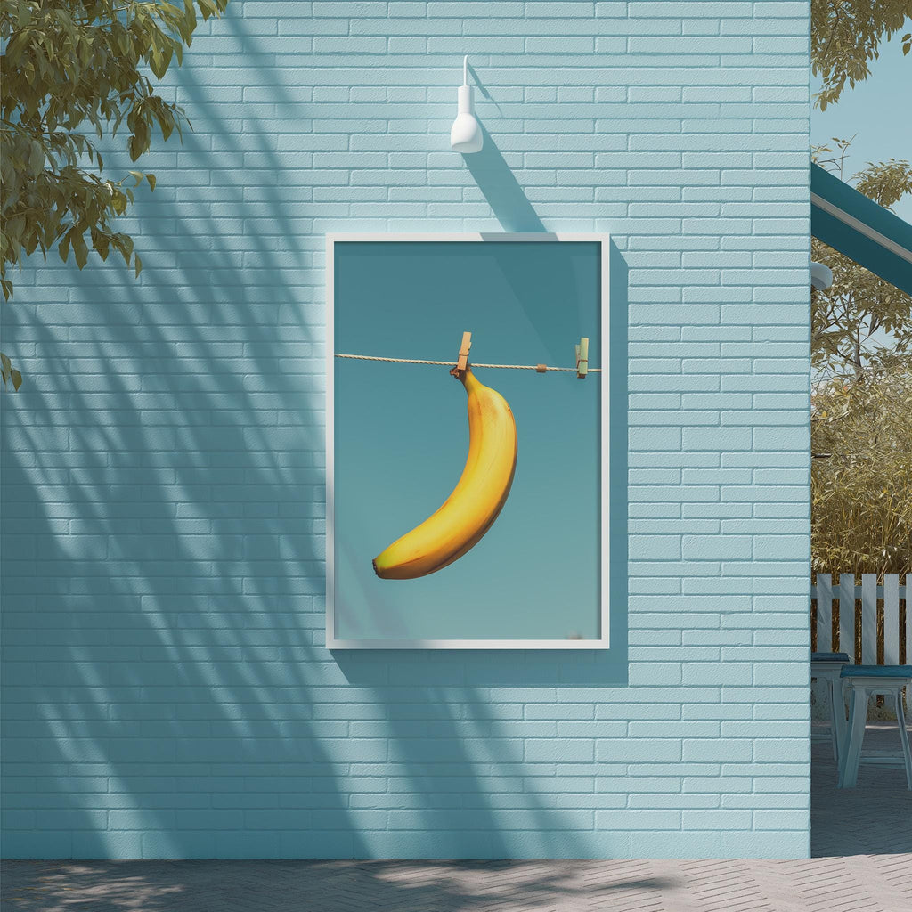 A framed image of a banana hanging on a clothesline is displayed on a blue brick wall.