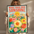 A woman is holding up a framed poster with a vibrant illustration of flowers and a sun.
