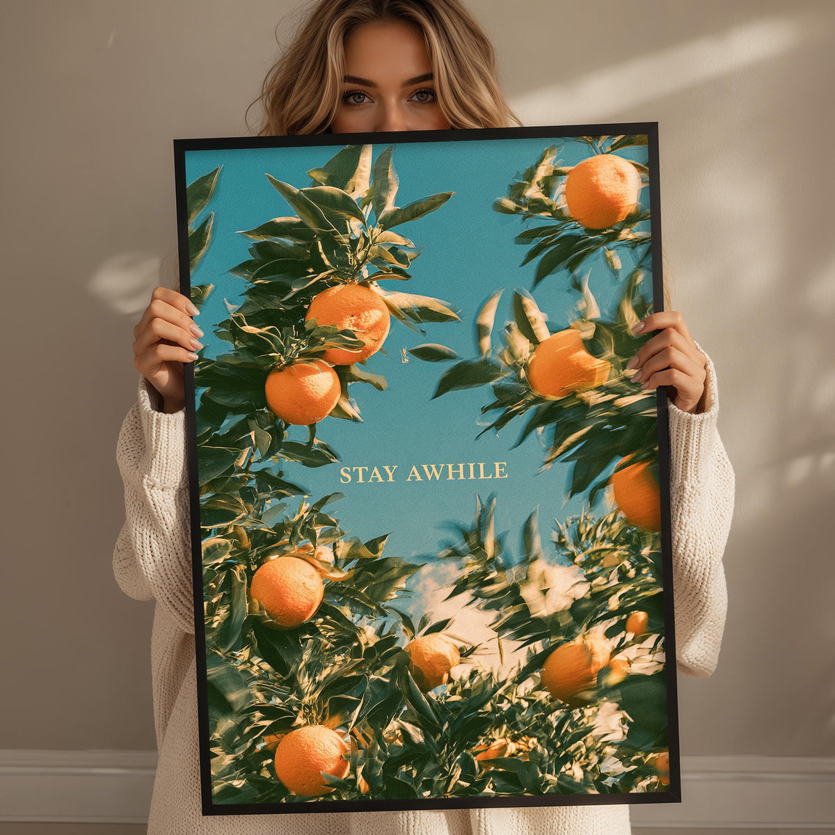 A woman holds up a framed poster with a vibrant image of orange trees and the text "STAY AWHILE" against a blue sky background.
