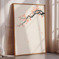 A large framed print depicting a branch with pink flowers is leaning against a white wall in a room with a window.
