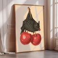 a framed illustration of a bat perched on two red cherries.