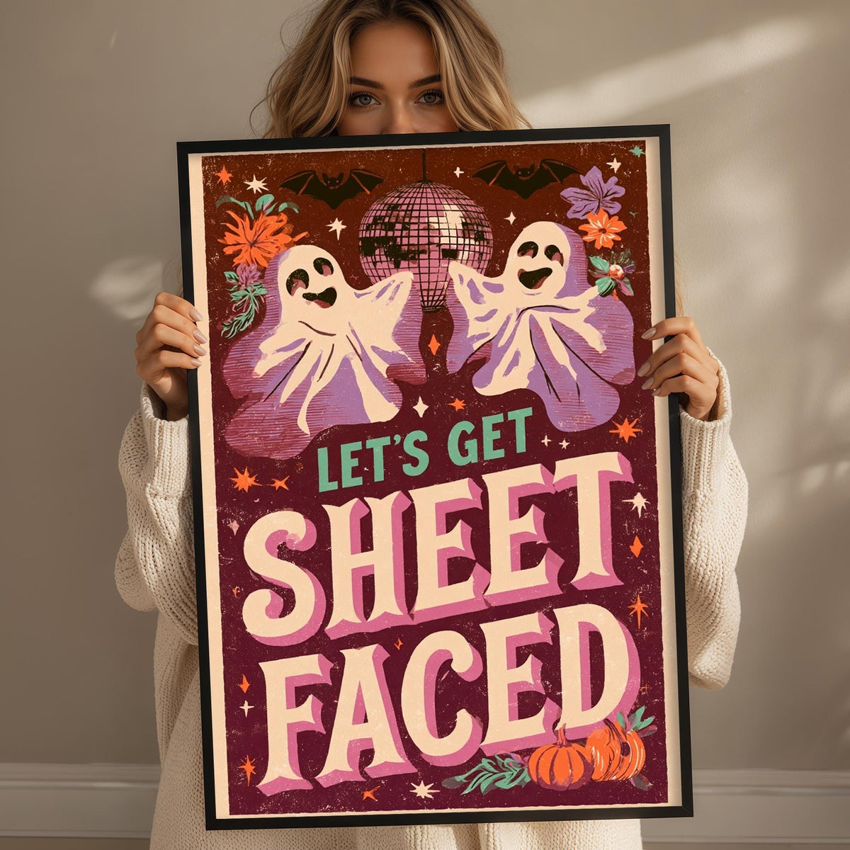 A woman is holding up a framed poster with a Halloween-themed design, including ghostly figures, a disco ball, and the text "Let's Get Sheetfaced".