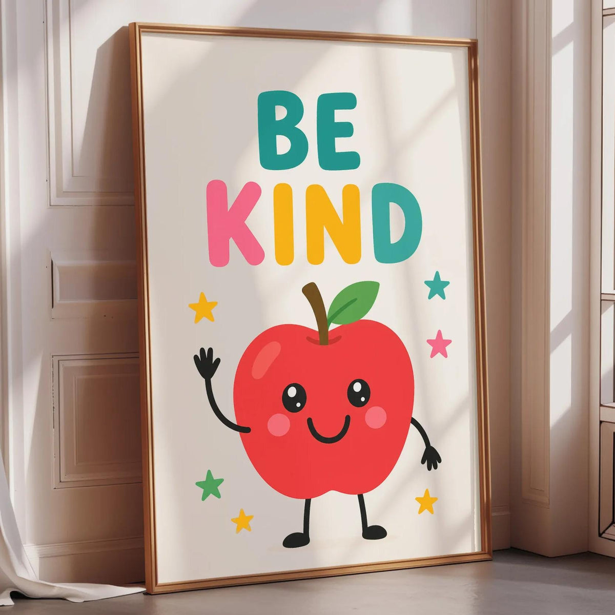 A framed poster with a red apple character and the text "BE KIND" on it, placed on a wooden floor near a window.