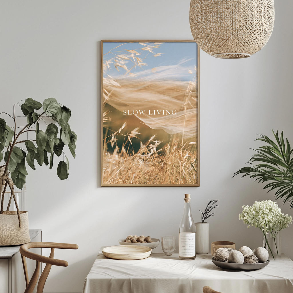 a dining table with a bowl of fruit, a bottle, and a vase of flowers, along with a potted plant and a hanging light fixture. Above the table, there is a framed picture of a field with a blue sky and clouds.