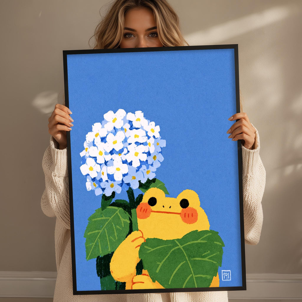 A woman holds up a framed illustration of a frog holding a bouquet of white flowers against a blue background.