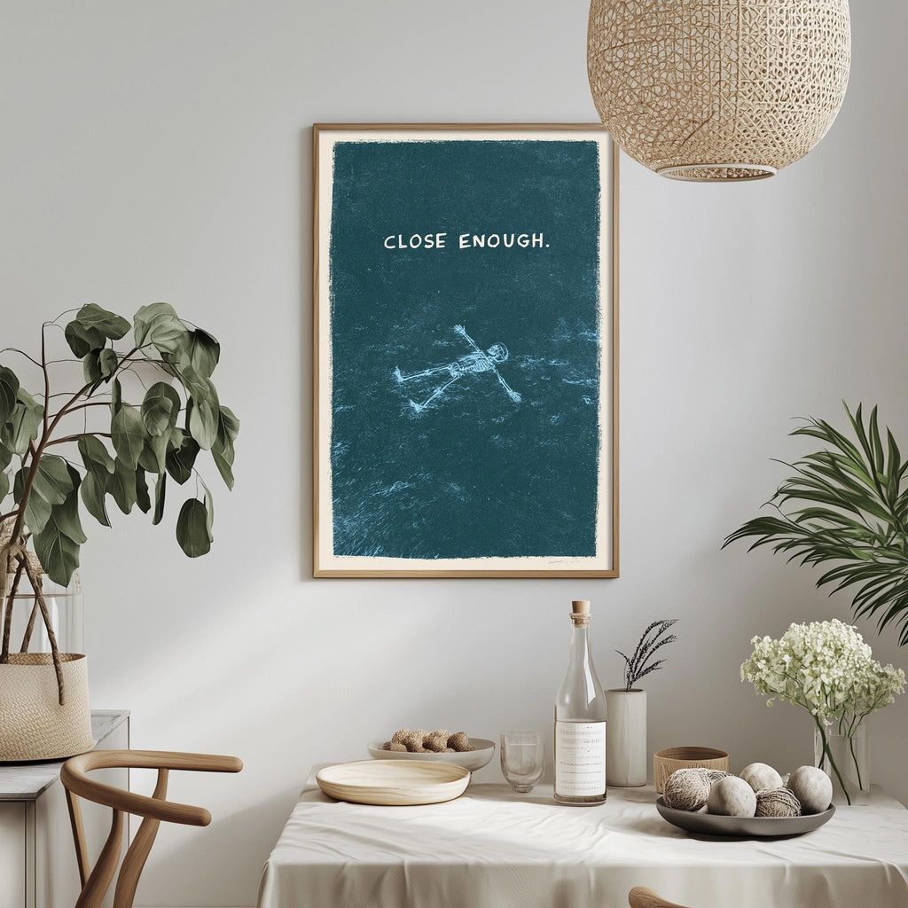 a framed poster with the text "CLOSE ENOUGH" hanging on a wall above a dining table with various items on it, including a bottle, a bowl, and a vase with flowers.