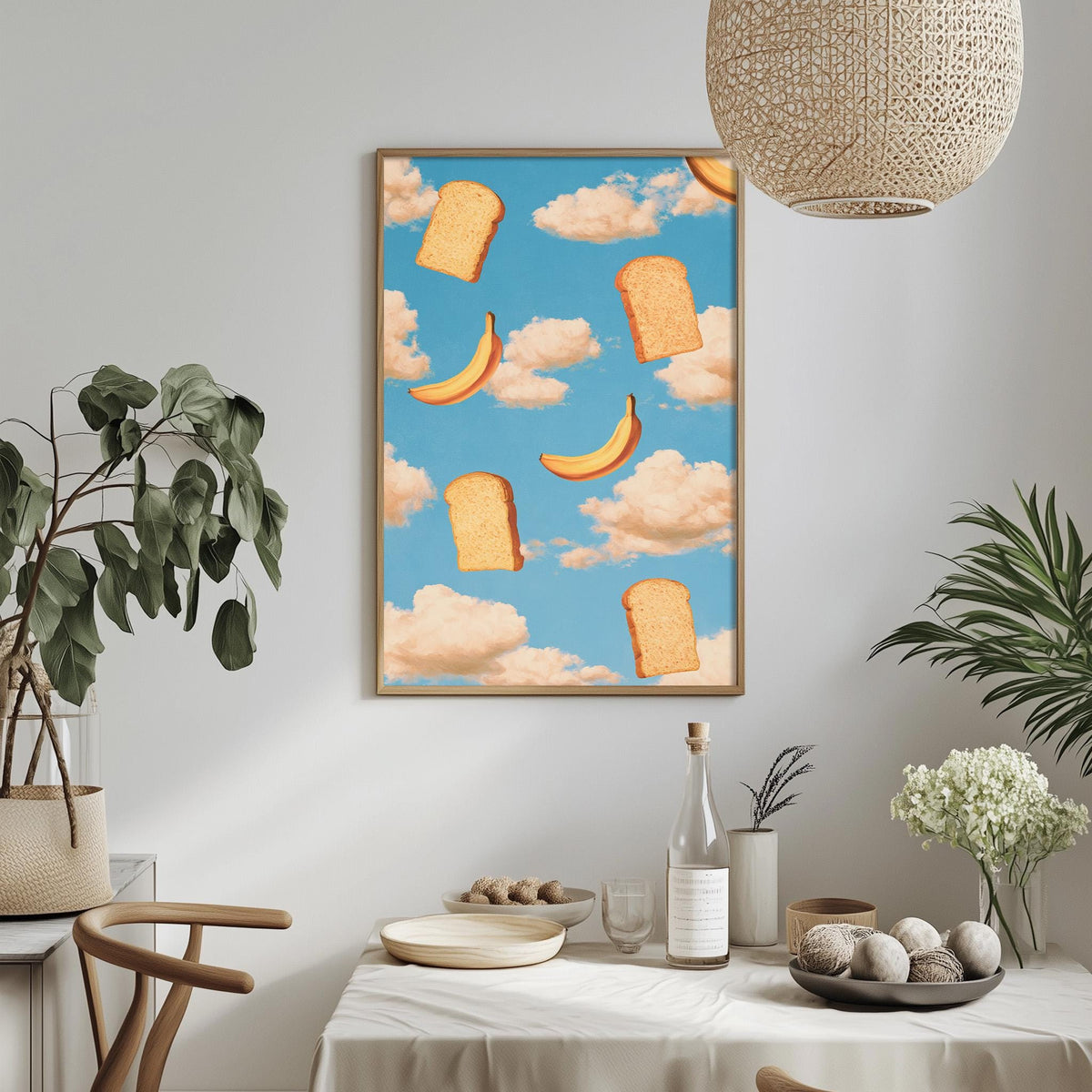 a dining table with a white tablecloth, a bowl of fruit, a bottle, and a vase with flowers. Above the table, there is a framed print of bread and bananas against a blue sky with clouds.