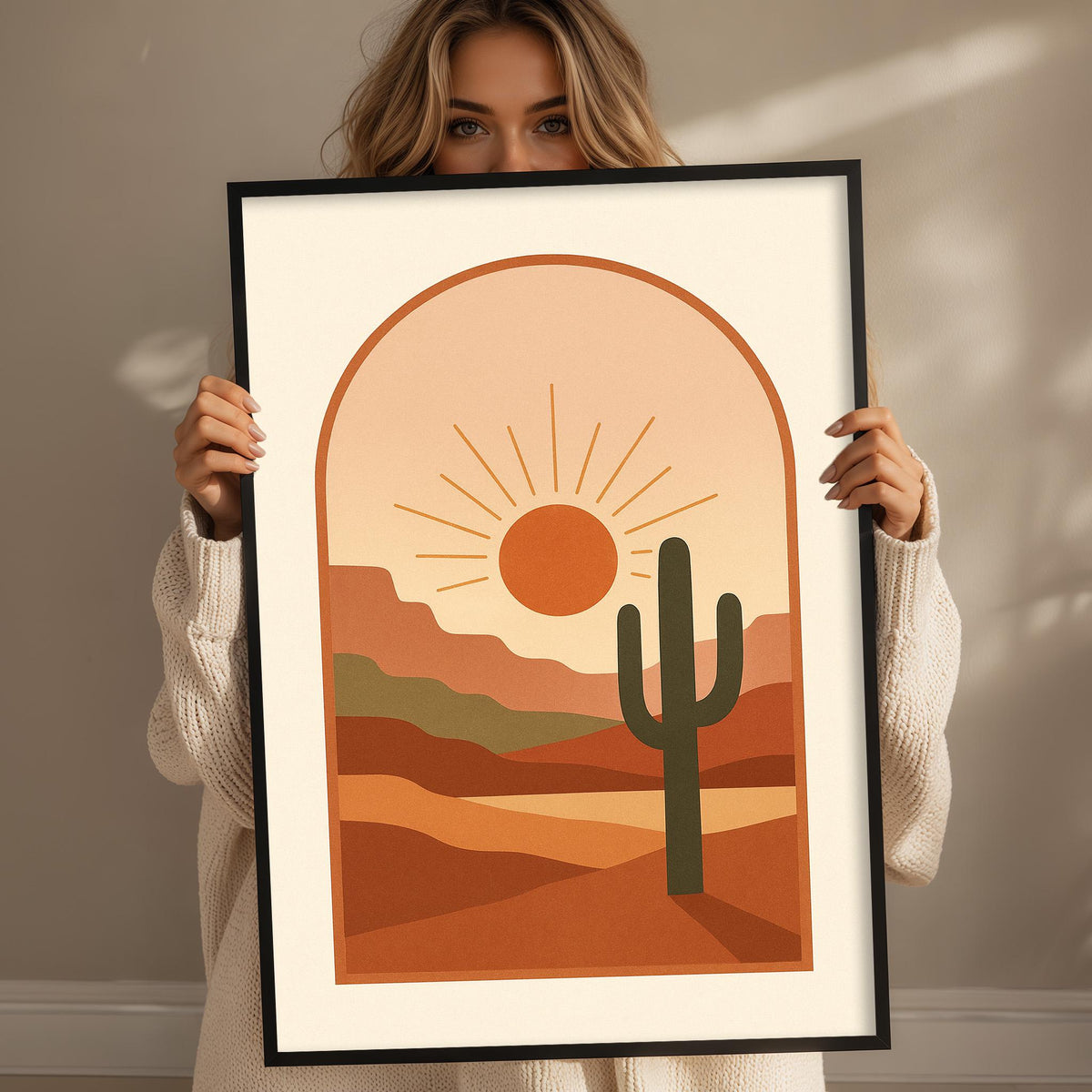 A woman is holding up a framed print of a desert landscape with a cactus in the foreground.