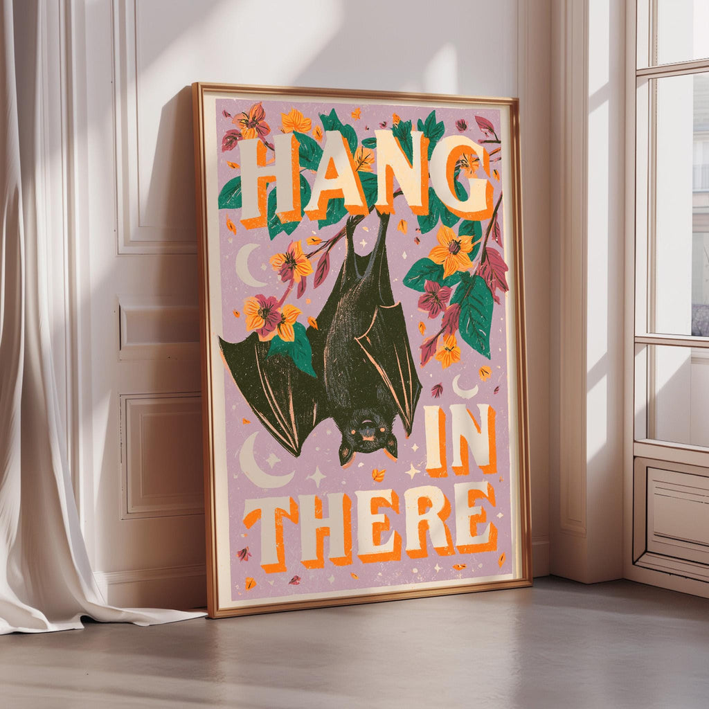 A framed poster with a bat illustration and the text "HANG IN THERE" on it, placed on a wooden floor next to a window.