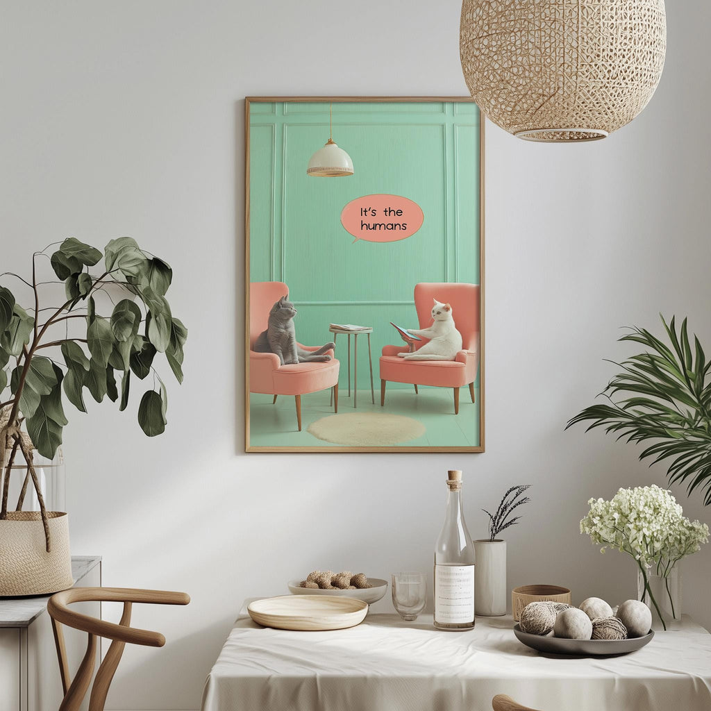 a framed poster of two cats sitting in a living room, with one cat sitting on a pink chair and the other on a red chair, and the text "It's the humans" written above them.