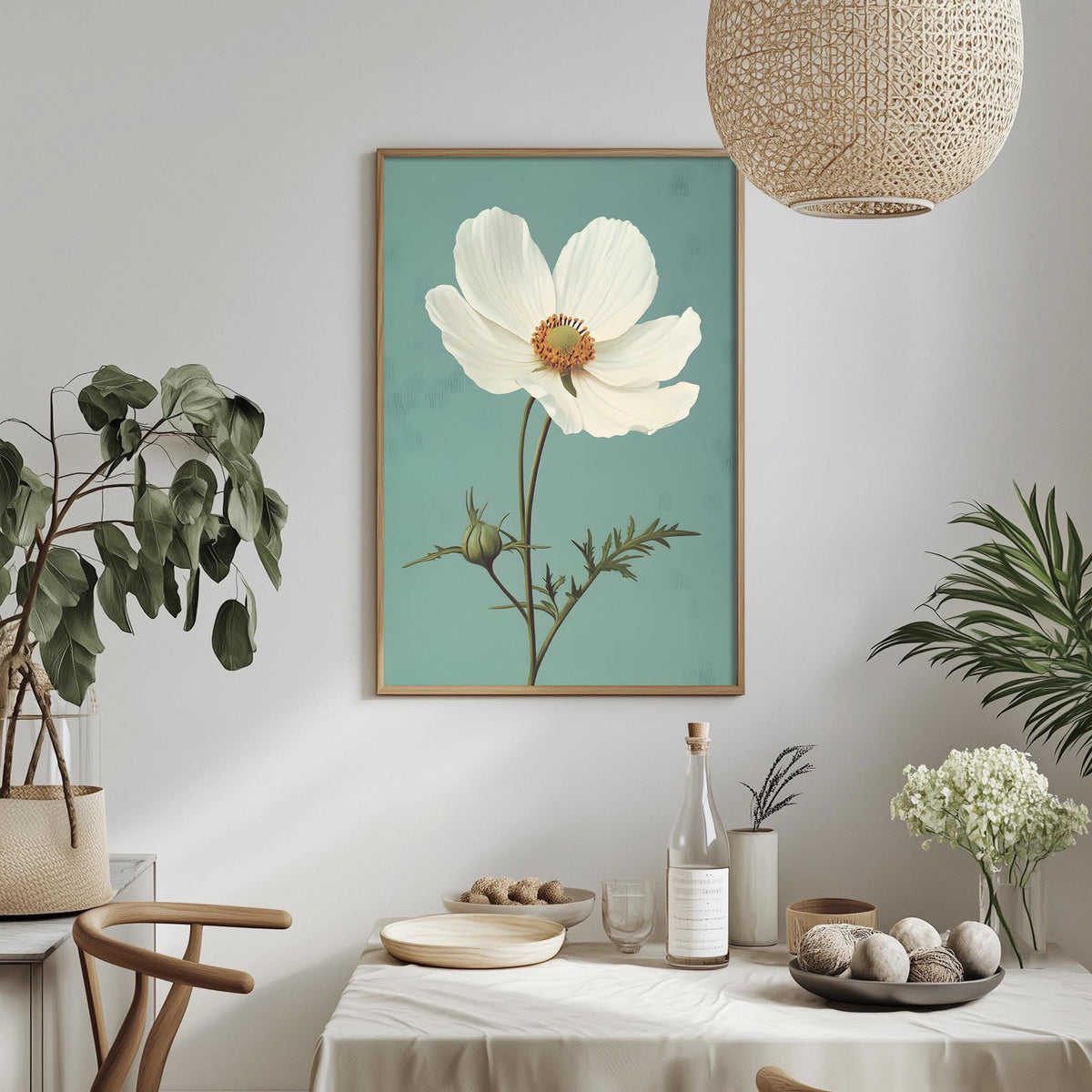 a dining table with a white tablecloth, a bowl of fruit, a bottle, and a vase with flowers. Above the table, there is a framed picture of a white flower with a green stem. The table is set with a plate, a wine glass, and a small bowl. The room has a minimalist and modern design, with a woven light fixture hanging above the table.