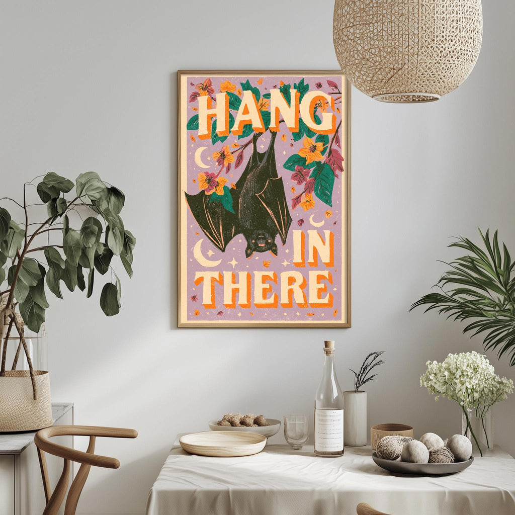 a framed poster with a bat illustration and the text "HANG IN THERE" hanging on a wall above a dining table with various items on it.