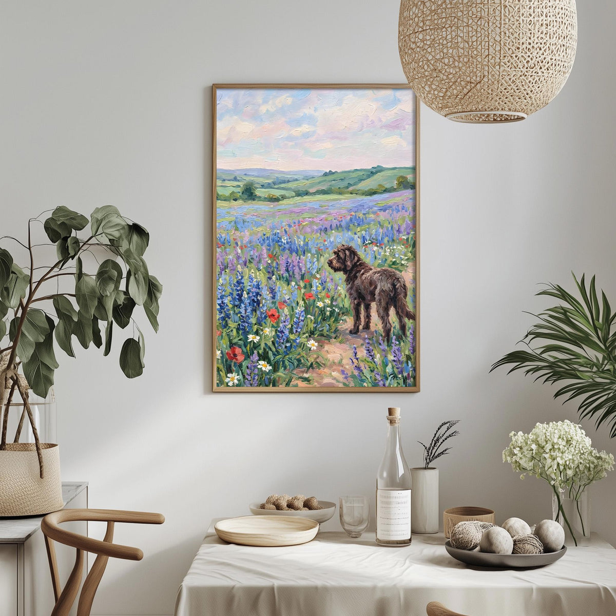 a painting of a dog standing in a field of flowers, with a serene landscape in the background. The painting is hung on a wall above a dining table, which has various items on it, including a bottle, a bowl, and a vase. The table is set with a white tablecloth and there are some plants and flowers in the room, creating a cozy and inviting atmosphere.