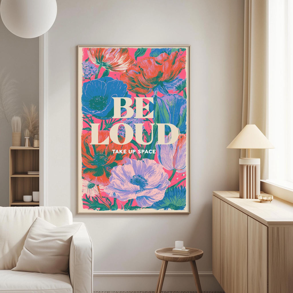 a large, colorful poster with the text "BE LOUD TAKE UP SPACE" displayed on it. The poster is hanging on a wall in a living room setting, with a white couch and a small table nearby.