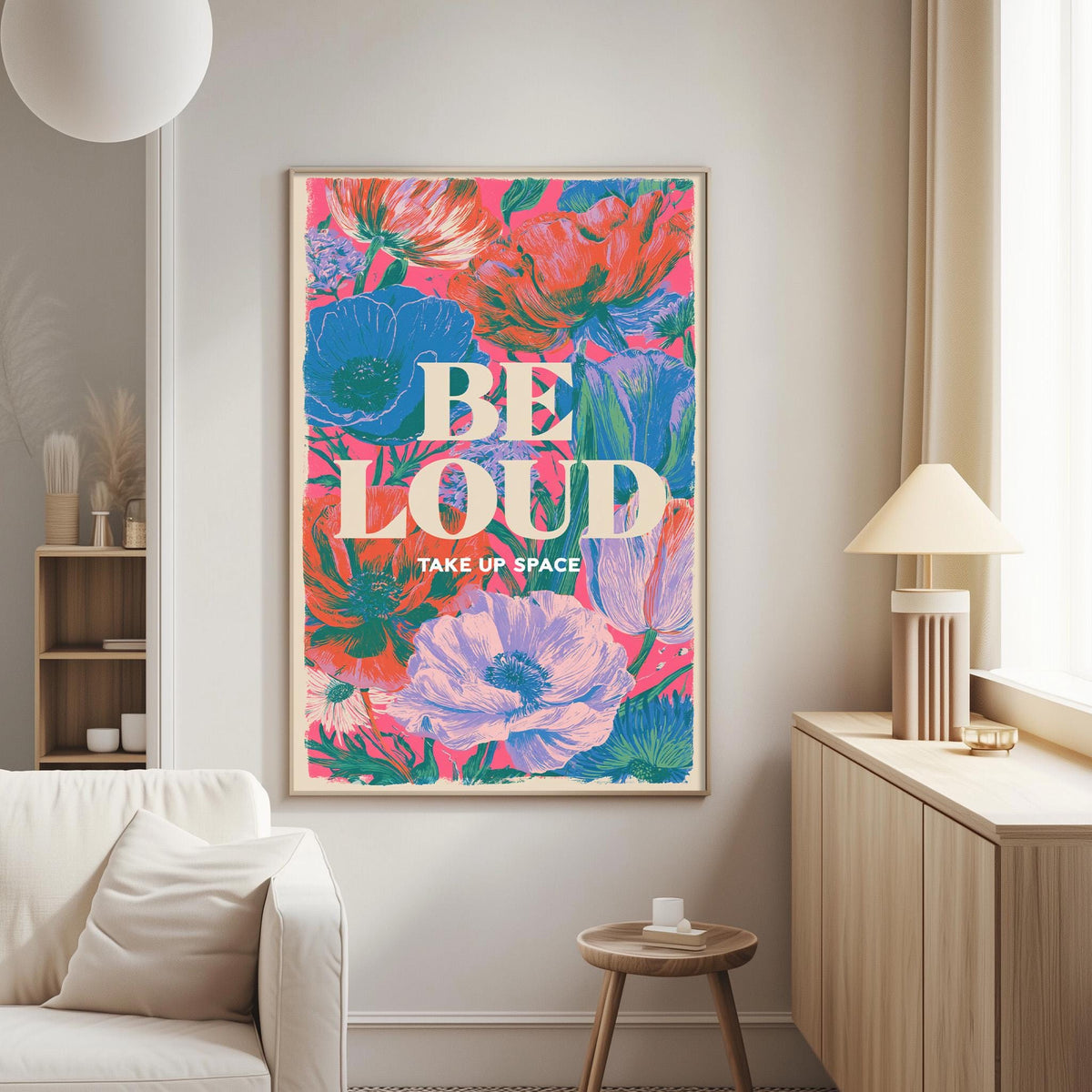 a large, colorful poster with the text "BE LOUD TAKE UP SPACE" displayed on it. The poster is hanging on a wall in a living room setting, with a white couch and a small table nearby.