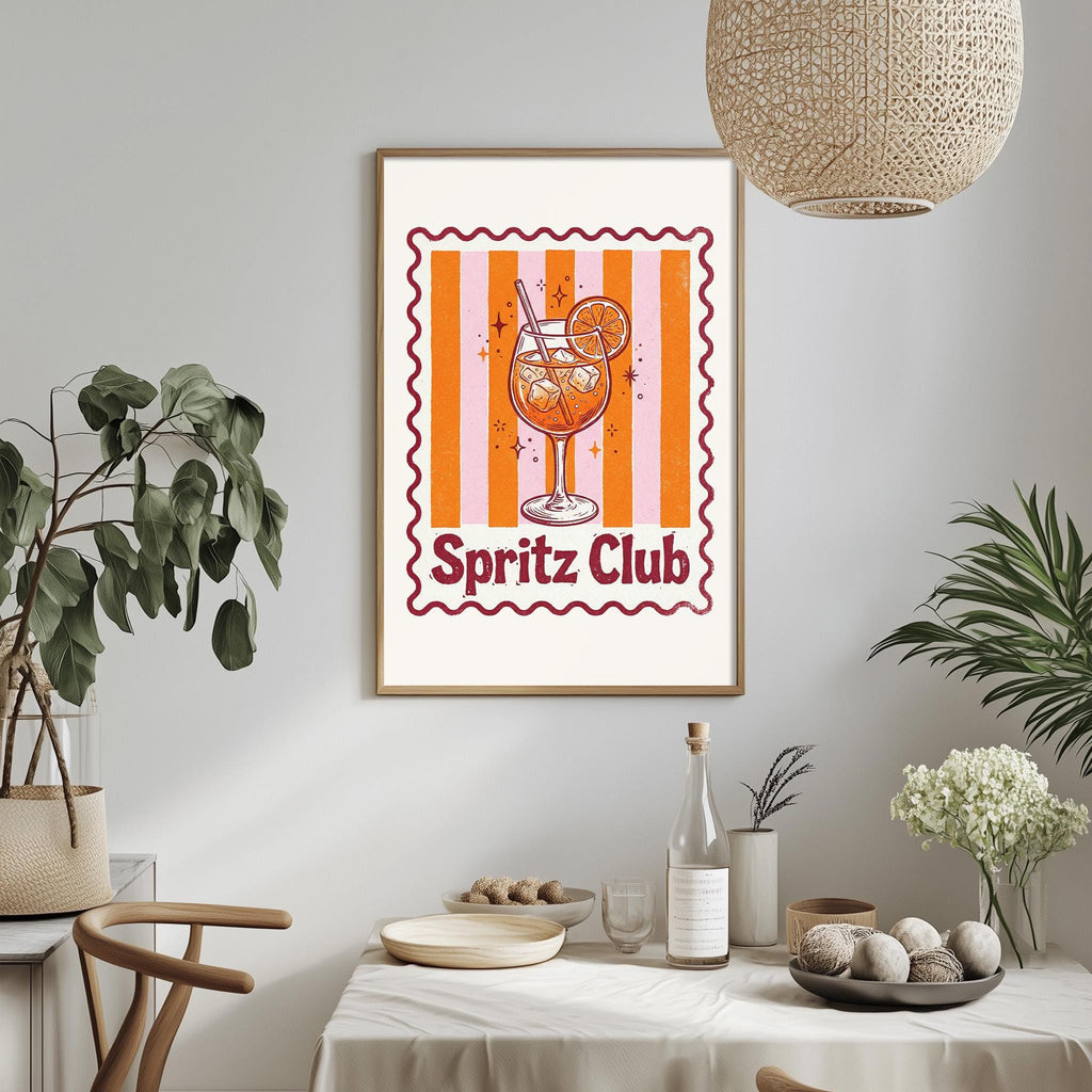 a framed poster of a glass of orange juice with the text "Spritz Club" on it, hanging on a wall above a dining table with various items on it.