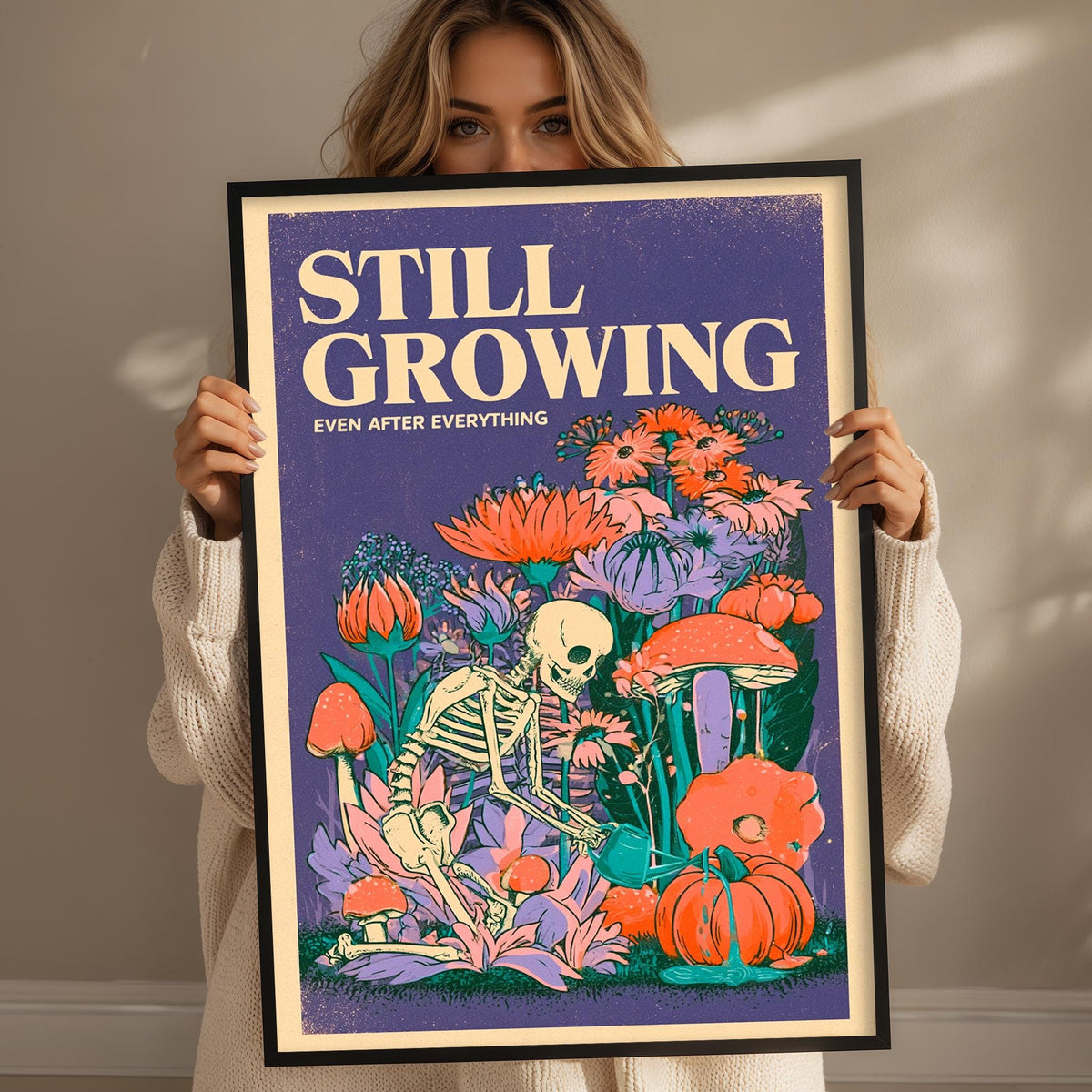 A woman is holding up a framed poster with a colorful illustration of a skeleton surrounded by flowers and mushrooms.