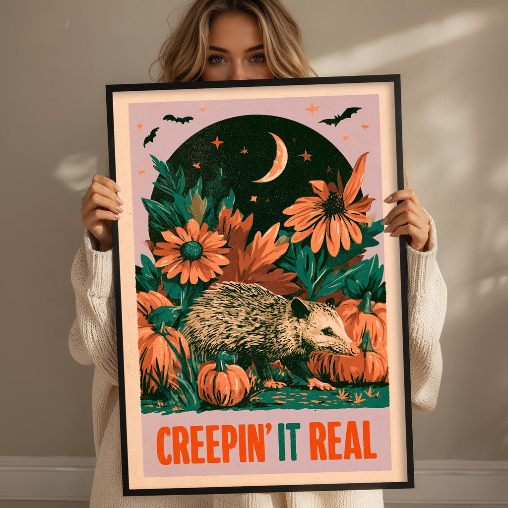 A woman is holding up a framed poster with a Halloween-themed illustration. The poster features a hedgehog surrounded by pumpkins and flowers, with a crescent moon and stars in the background. The woman is wearing a white sweater and is holding the poster up to her face.