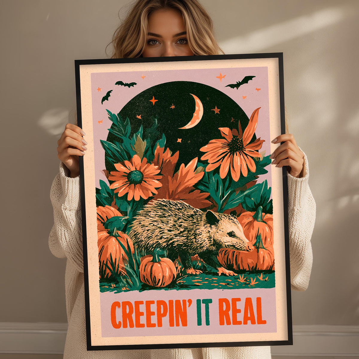 A woman is holding up a framed poster with a Halloween-themed illustration. The poster features a hedgehog surrounded by pumpkins and flowers, with a crescent moon and stars in the background. The woman is wearing a white sweater and is holding the poster up to her face.