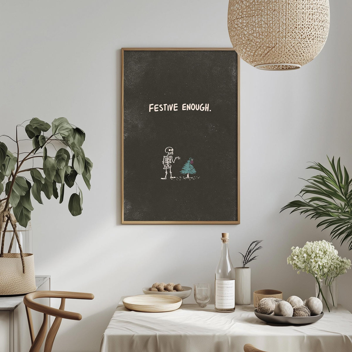 a dining table with a white tablecloth, a bowl of fruit, a bottle, and a vase with flowers. Above the table, there is a framed poster with the text "FESTIVE ENOUGH" and a drawing of a skeleton holding a Christmas tree.
