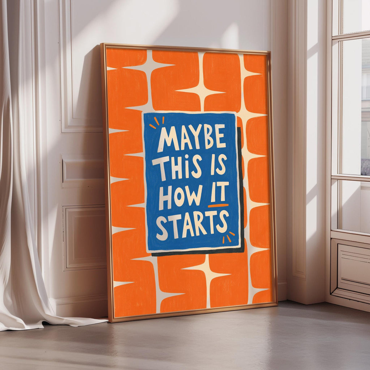 a framed poster with the text "MAYBE THIS IS HOW IT STARTS" on it, placed on a wooden floor next to a window.